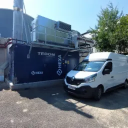 Kogeneracja TEDOM HEXA z białym vanem dostawczym Renault na tle budynków przemysłowych i drzew w słoneczny dzień.