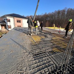 Firma budowlana Domy Raz Dwa - Wylewanie betonu na strop domu jednorodzinnego z widoczną siatką zbrojeniową i pracownikami w tle, słoneczny dzień.