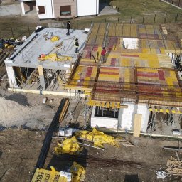 Firma budowlana Domy Raz Dwa - Budowa domu jednorodzinnego z widocznymi etapami: wylany betonowy strop i przygotowania do zalania stropu z szalunkami i zbrojeniami. Widok z góry na plac budowy i okoliczne tereny.