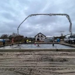 Wylewanie betonu na przygotowany fundament pod budowę domu jednorodzinnego; widoczny betonowóz z pompą, pracownicy w odblaskowych kamizelkach i szalunek.