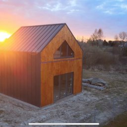 Firma budowlana Domy Raz Dwa - Nowoczesny dom murowany o prostej bryle z drewnianą elewacją i metalowym dachem, uchwycony o zachodzie słońca, z widocznymi elementami placu budowy.