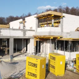 Wznoszenie ścian domu z bloczków Ytong, widoczne podpory szalunkowe, częściowo zabetonowane nadproża i przygotowane pakiety bloczków na placu budowy.