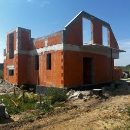 Firma budowlana Domy Raz Dwa - Budowany dom jednorodzinny w stanie surowym otwartym, z murami z czerwonych pustaków ceramicznych i betonowymi nadprożami. Widoczne otwory okienne, drzwiowe oraz konstrukcja dachu poddasza.