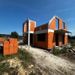 Firma budowlana Domy Raz Dwa - Wzniesione ściany domu jednorodzinnego z pomarańczowych pustaków ceramicznych, z betonowymi nadprożami i słupami, oraz małym zadaszeniem wejścia, na tle błękitnego nieba.