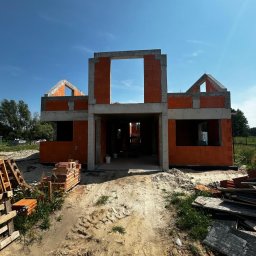 Firma budowlana Domy Raz Dwa - Częściowo zbudowany dom z pomarańczowych pustaków ceramicznych i betonowych nadproży, z widocznymi otworami okiennymi i wejściem, na piaszczystym placu budowy w słoneczny dzień.