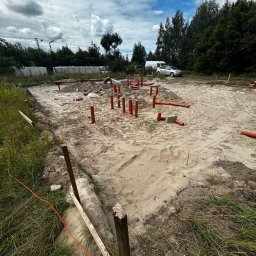 Firma budowlana Domy Raz Dwa - Plac budowy z przygotowanym wykopem pod fundament, gdzie z piaszczystego podłoża wystają liczne pomarańczowe rury instalacji wodno-kanalizacyjnej, otoczony trawą i drzewami.