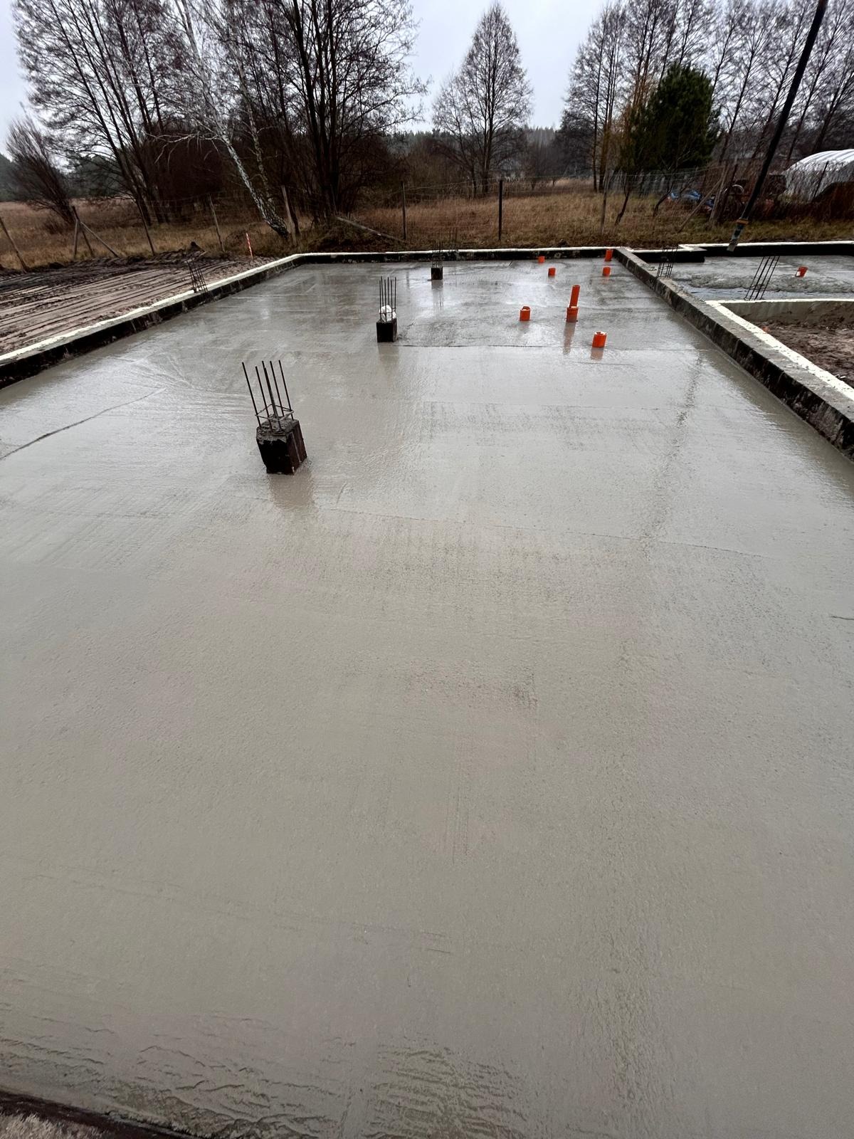 Wylany, świeży betonowy fundament z wystającymi prętami zbrojeniowymi i pomarańczowymi rurami instalacyjnymi na obrzeżach działki budowlanej.