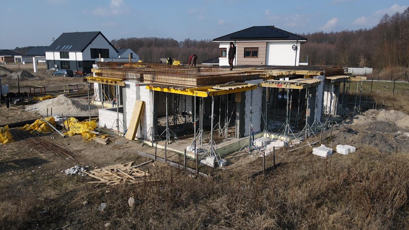 Budowa domu jednorodzinnego w stanie surowym otwartym: widoczne ściany z bloczków, szalunki stropowe wsparte na stemplach, zbrojenie na stropie, w tle inne domy w zabudowie jednorodzinnej.