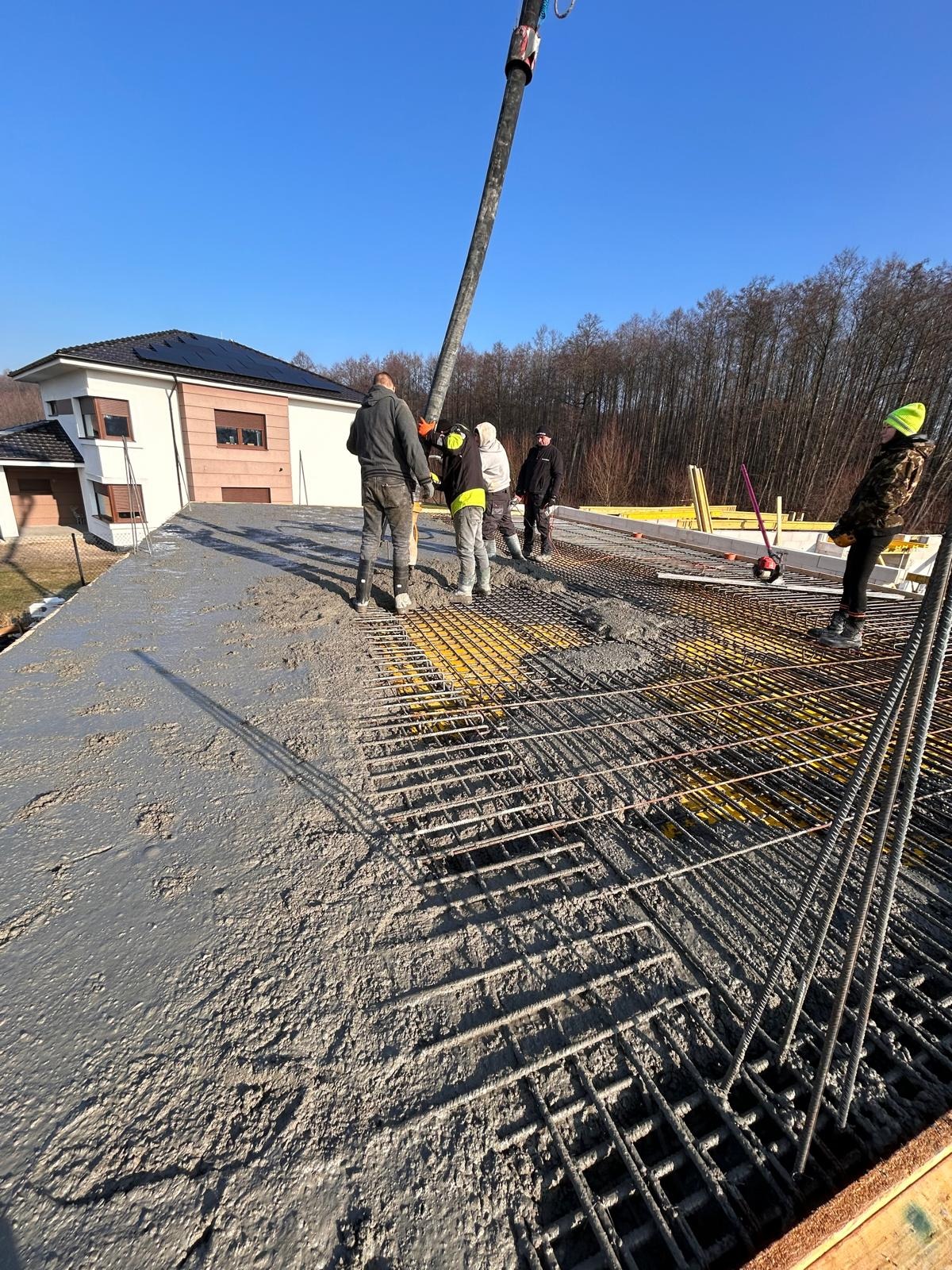 Wylewanie betonu na strop domu jednorodzinnego z widoczną siatką zbrojeniową i pracownikami w tle, słoneczny dzień.