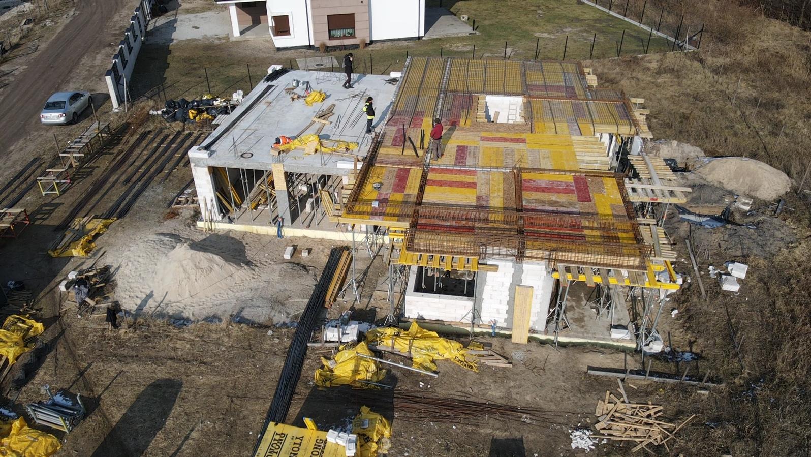 Budowa domu jednorodzinnego z widocznymi etapami: wylany betonowy strop i przygotowania do zalania stropu z szalunkami i zbrojeniami. Widok z góry na plac budowy i okoliczne tereny.