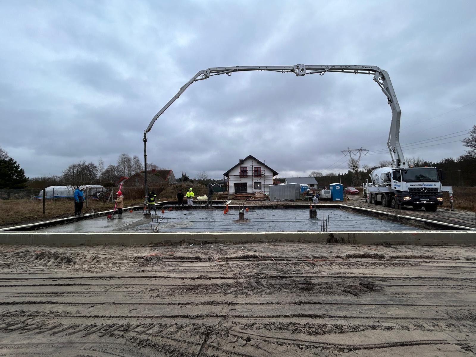 Wylewanie betonu na przygotowany fundament pod budowę domu jednorodzinnego; widoczny betonowóz z pompą, pracownicy w odblaskowych kamizelkach i szalunek.