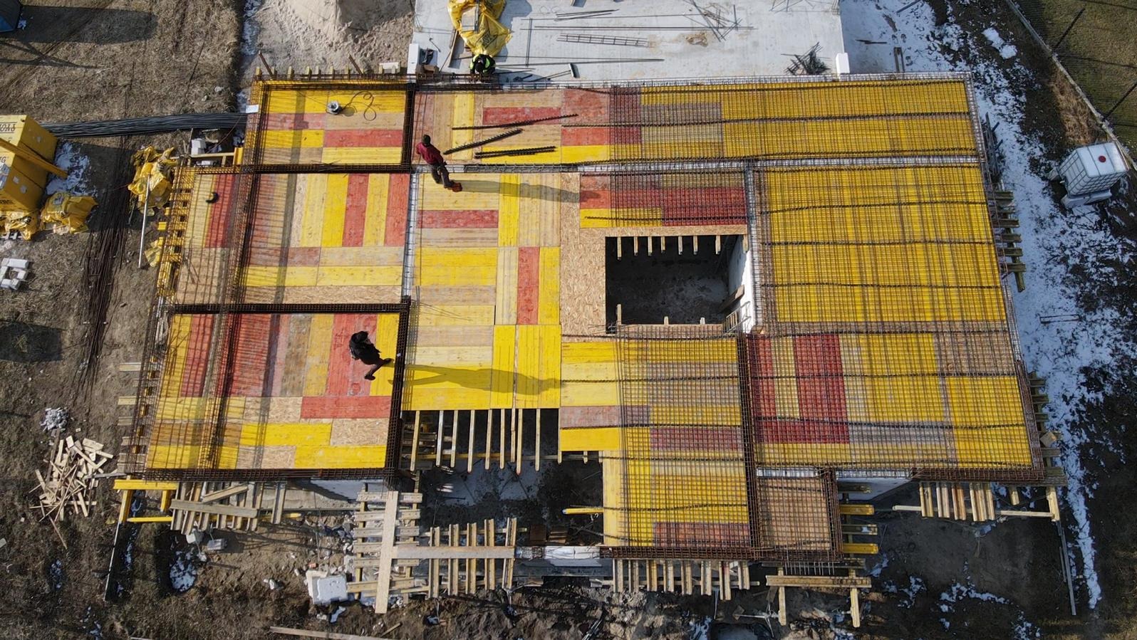 Widok z góry na plac budowy z przygotowanymi szalunkami i zbrojeniami fundamentów domu, z widocznymi pracownikami i elementami konstrukcyjnymi.