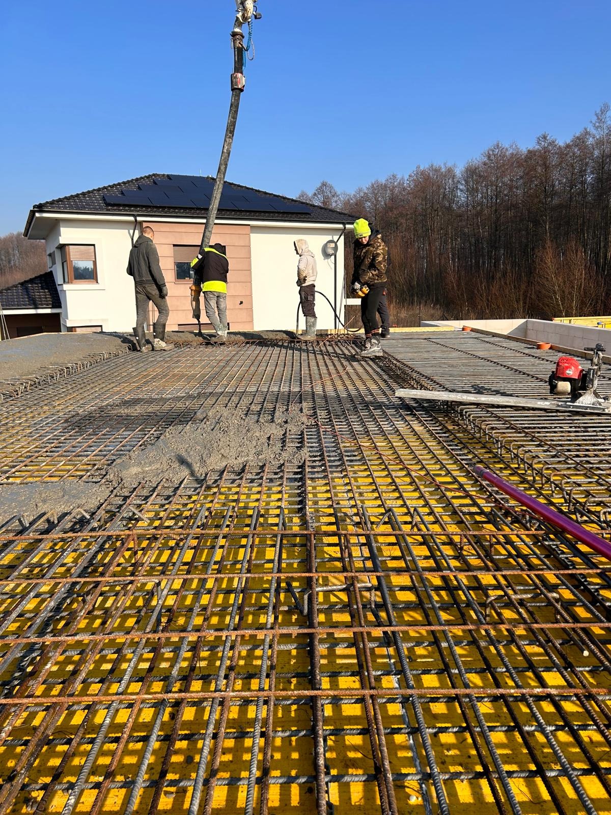 Wylewanie betonu na zbrojenie stropu domu jednorodzinnego z widocznymi deskami szalunkowymi, pracownikami i pompą do betonu w tle.