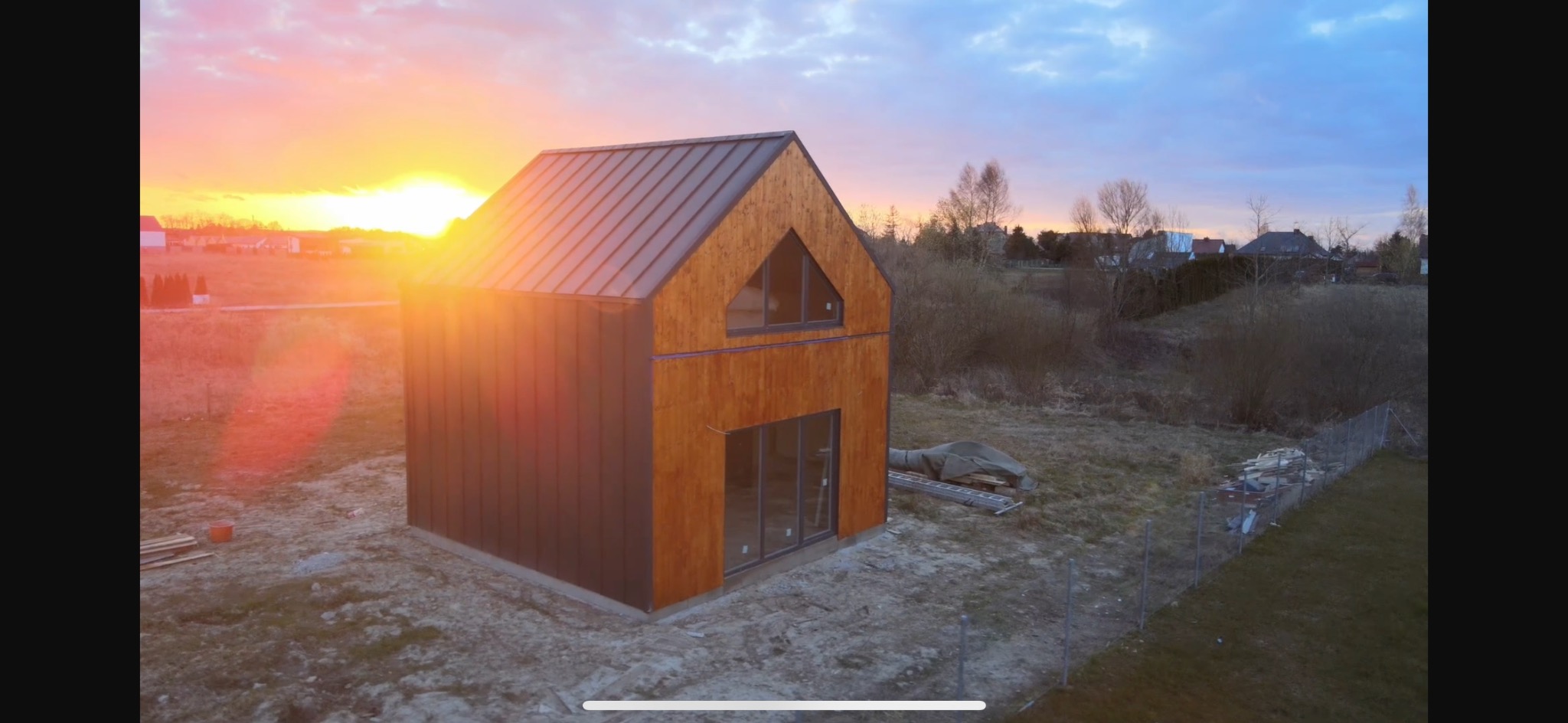 Nowoczesny dom murowany o prostej bryle z drewnianą elewacją i metalowym dachem, uchwycony o zachodzie słońca, z widocznymi elementami placu budowy.