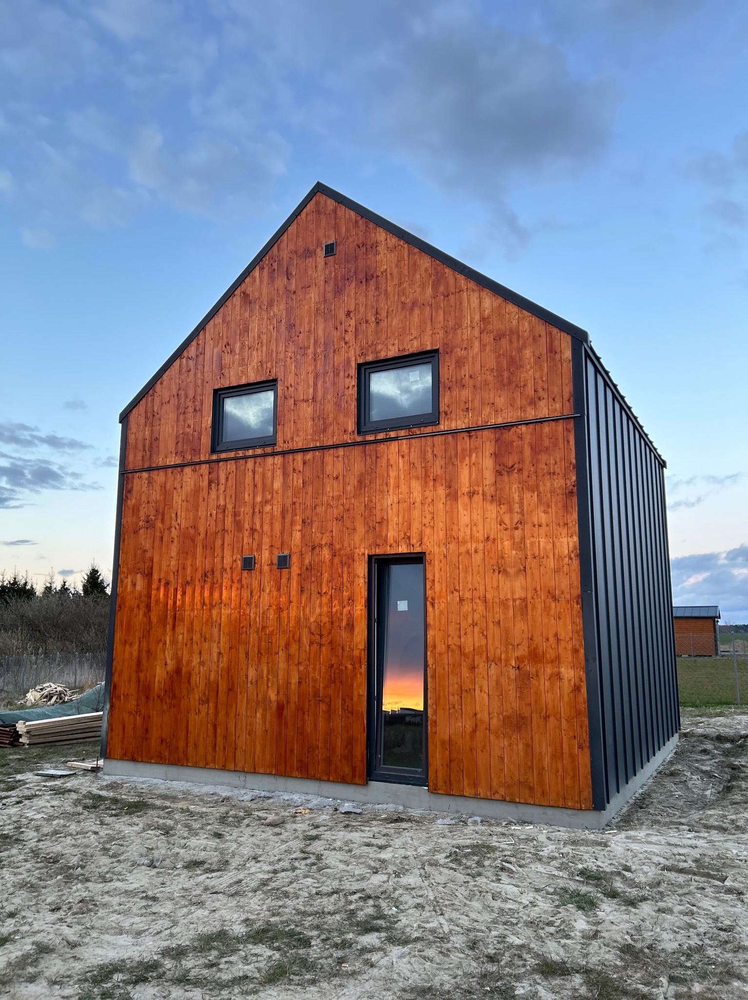 Nowoczesny dom z drewnianą elewacją i czarnymi akcentami, widoczne dwa okna na górnej kondygnacji i drzwi wejściowe z odbiciem zachodu słońca, teren wokół domu w trakcie prac wykończeniowych.