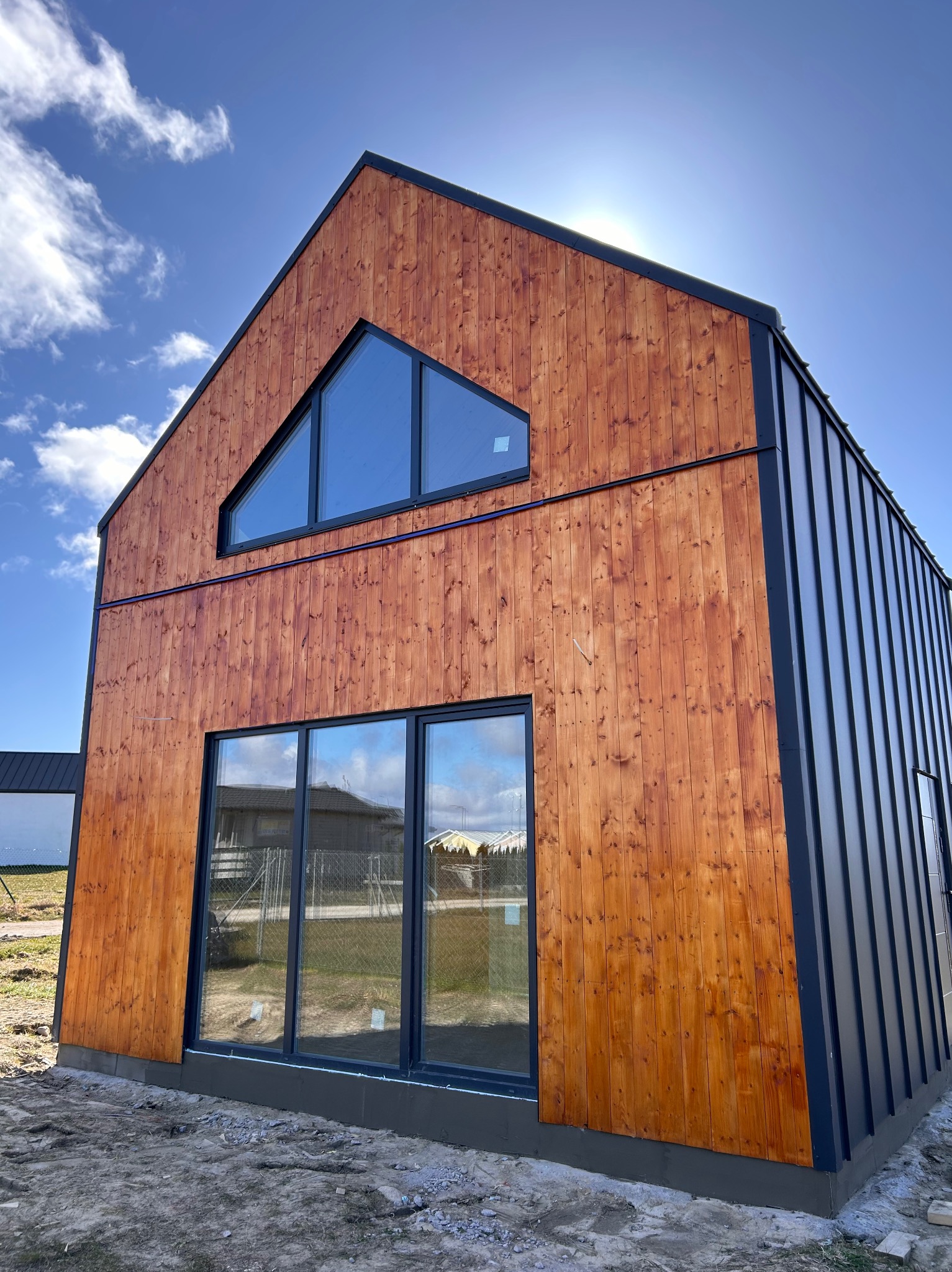 Nowoczesny dom z drewnianą elewacją i trójkątnym oknem na szczycie, metalowe wykończenia, widok z dołu na tle błękitnego nieba z chmurami.