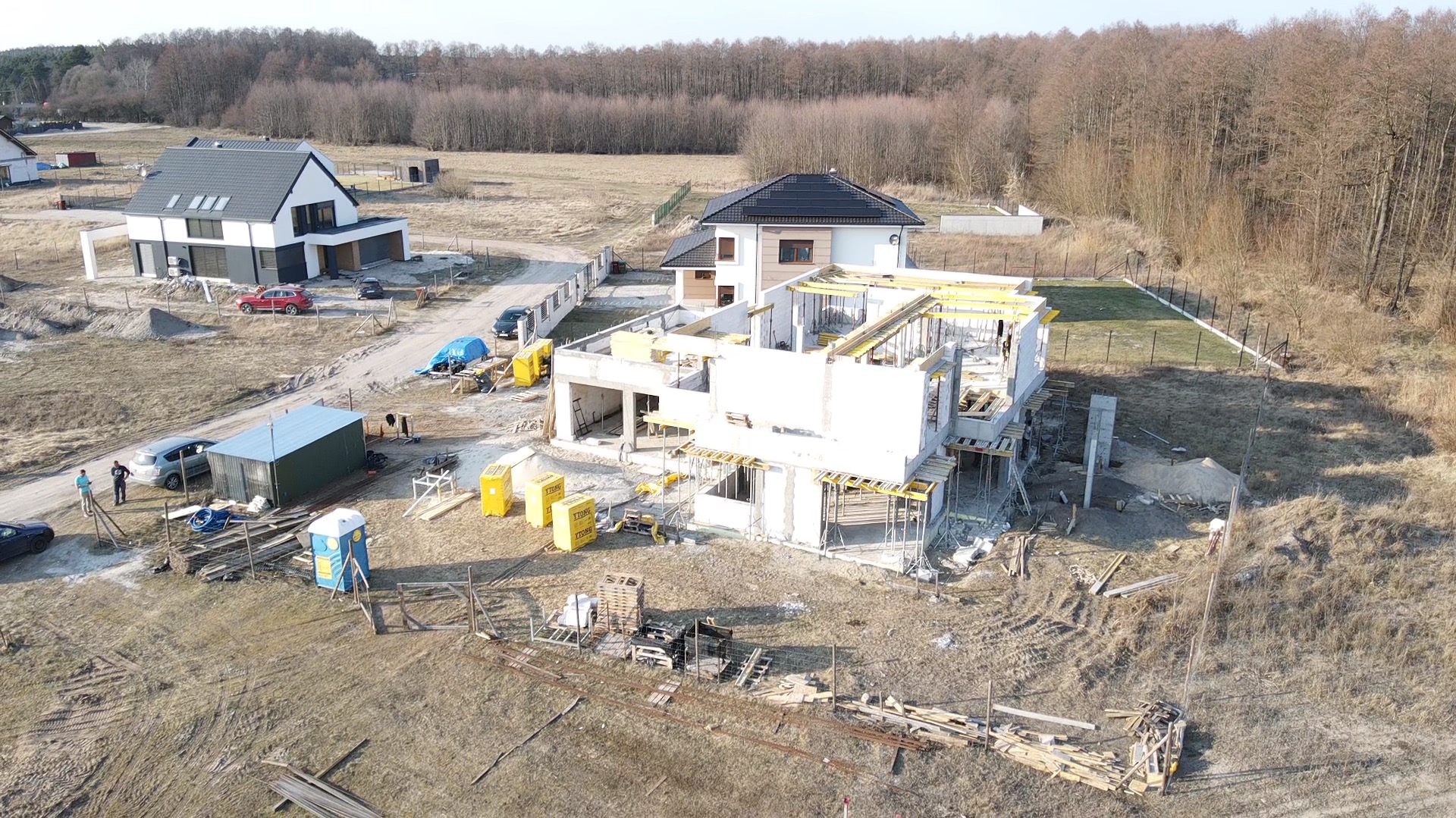 Budowa domu w stanie surowym otwartym, widok z lotu ptaka na plac budowy z żółtymi bloczkami Ytong, szalunkami i dwoma ukończonymi domami w tle.