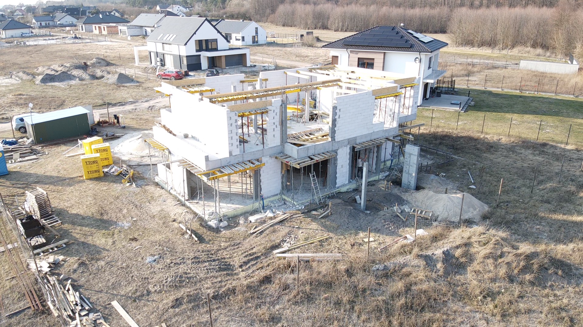 Wznoszenie dwukondygnacyjnego budynku z białych bloczków, widoczne szalunki i podpory, w tle zabudowa jednorodzinna i drzewa bezlistne.