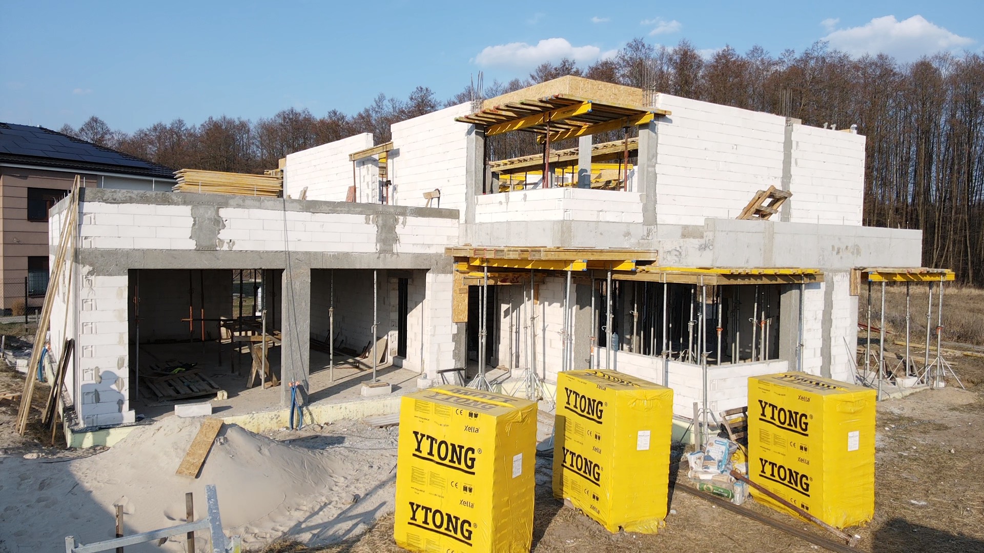 Wznoszenie ścian domu z bloczków Ytong, widoczne podpory szalunkowe, częściowo zabetonowane nadproża i przygotowane pakiety bloczków na placu budowy.
