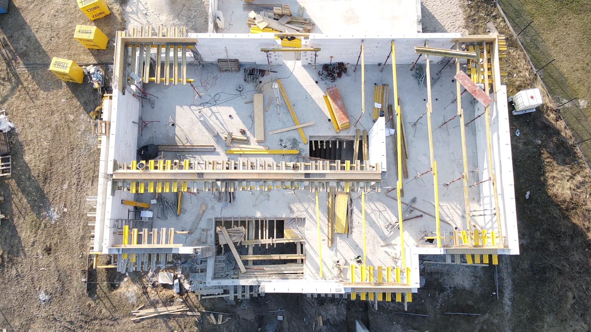 Widok z góry na plac budowy domu z białych bloczków, z żółtymi elementami szalunków i drewnianymi konstrukcjami, w tle trawa i ogrodzenie.