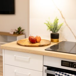 Joanna Lar - Plena Invest - Jasna kuchnia z drewnianym blatem, na którym leżą trzy czerwone jabłka na drewnianej tacy, obok stoi roślina w czarno-złotej doniczce, w tle biała ściana z marmurowym wzorem i telewizor