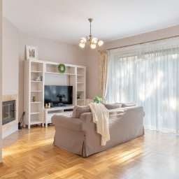 Joanna Lar - Plena Invest - Jasne, przestronne wnętrze salonu z drewnianymi schodami, parkietem w jodełkę, beżową sofą z narzutą, białymi regałami, telewizorem, kominkiem i zasłonami w kwiatowy wzór, oświetlone światłem...