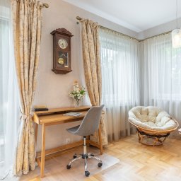 Joanna Lar - Plena Invest - Jasne wnętrze pokoju z drewnianym biurkiem, szarym krzesłem na kółkach i okrągłym fotelem z poduszką, oświetlone naturalnym światłem wpadającym przez okna z delikatnymi, wzorzystymi zasłonami.