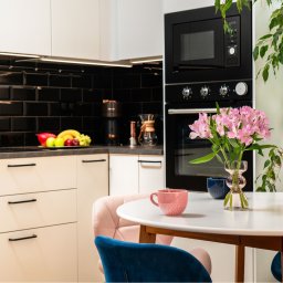 Joanna Lar - Plena Invest - Jasna kuchnia w stylu nowoczesnym z czarnymi płytkami, kwiatami na stole i owocami na blacie. Przytulne wnętrze. Okrągły stół z filiżankami i krzesłami.