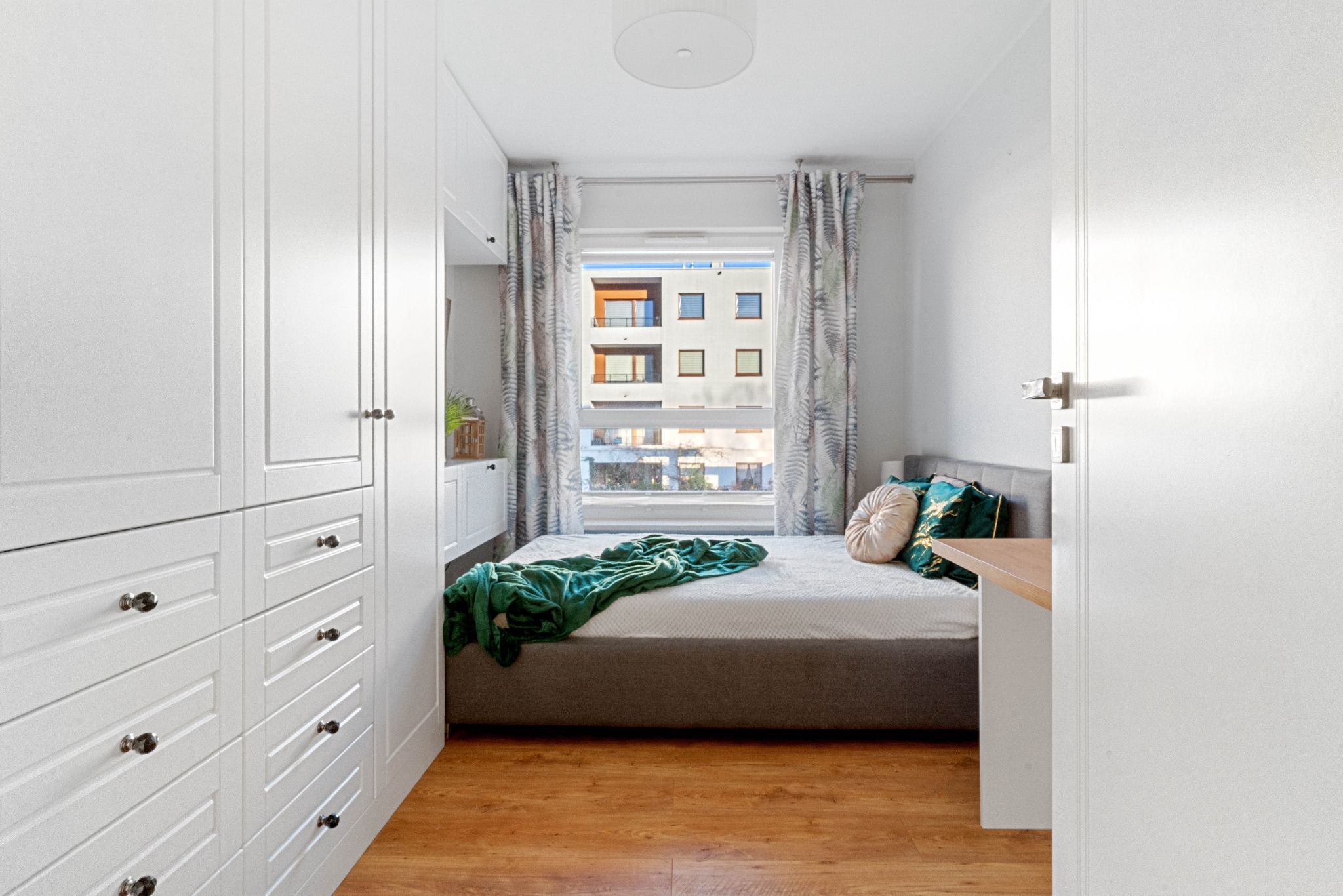 Kompaktowa sypialnia z dużym, białym, wbudowanym systemem szaf, łóżkiem z szarą ramą i zielonym pledem, widok z korytarza przez otwarte drzwi.