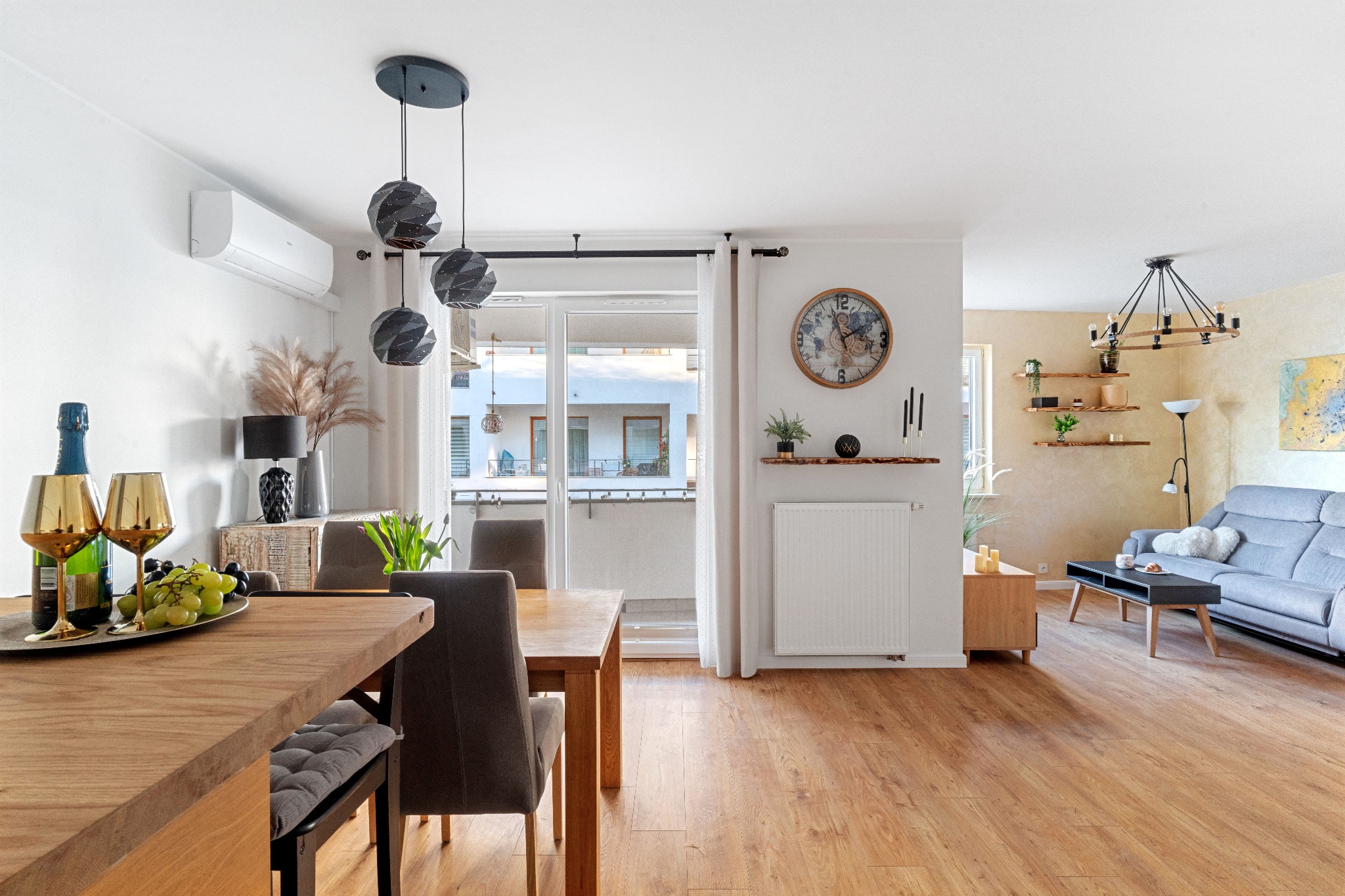 Jasne wnętrze mieszkania z drewnianą podłogą, stołem z owocami i szampanem, widok na balkon i salon z szarą sofą.