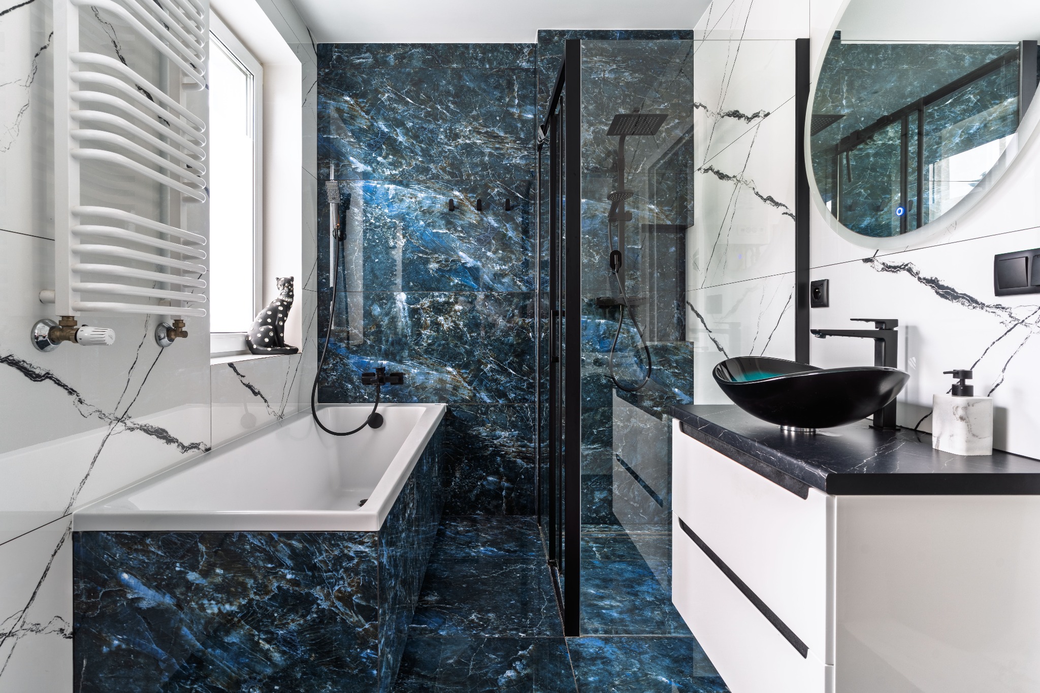 Nowoczesna łazienka z wanną obudowaną płytkami imitującymi niebieski marmur, czarną kabiną prysznicową, umywalką w kształcie misy i okrągłym lustrem z podświetleniem LED.