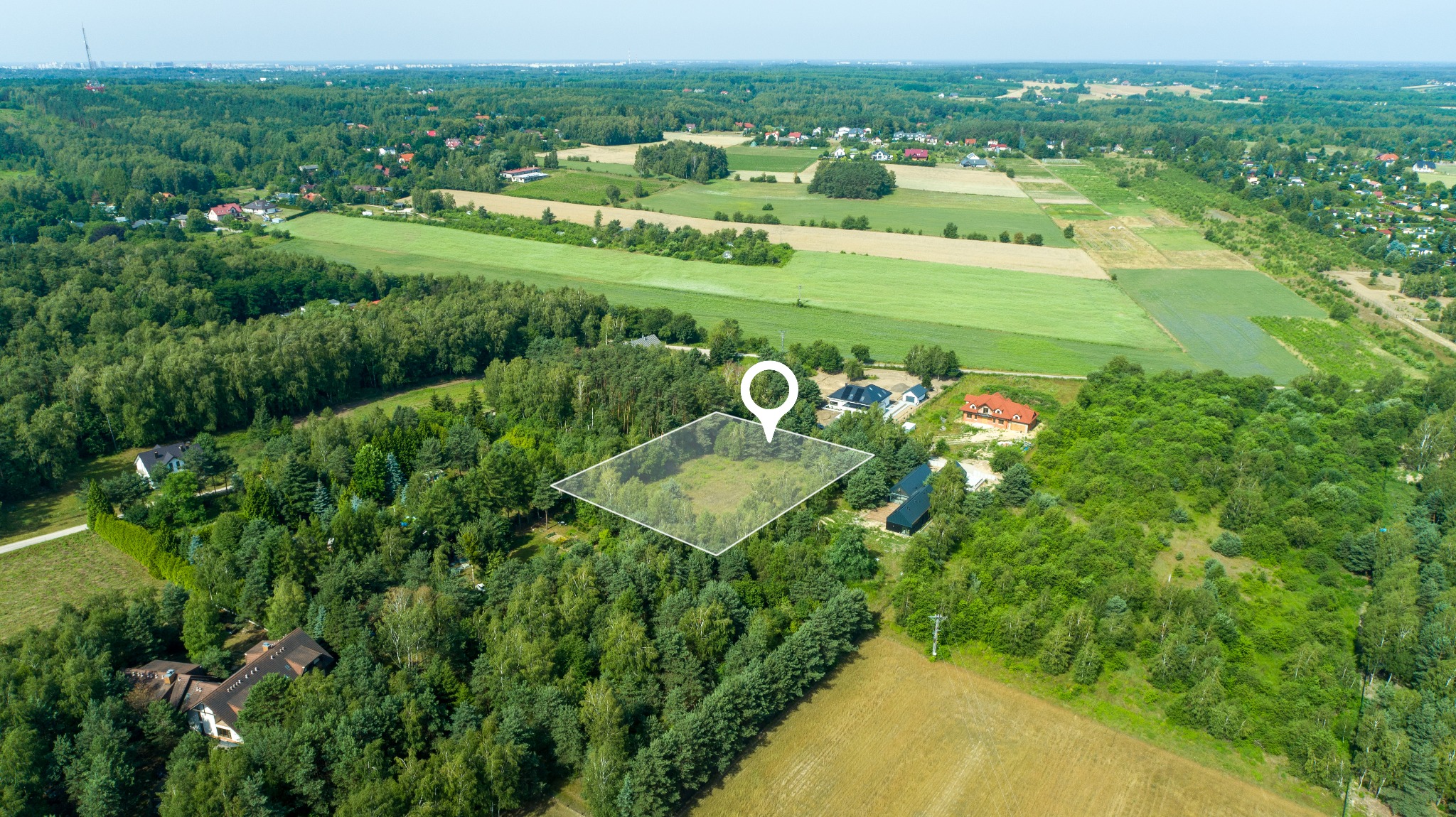 Działka budowlana z zaznaczonymi granicami i pinezką lokalizacyjną, widok z lotu ptaka na zalesiony teren pod zabudowę w otoczeniu domów jednorodzinnych i pól uprawnych.