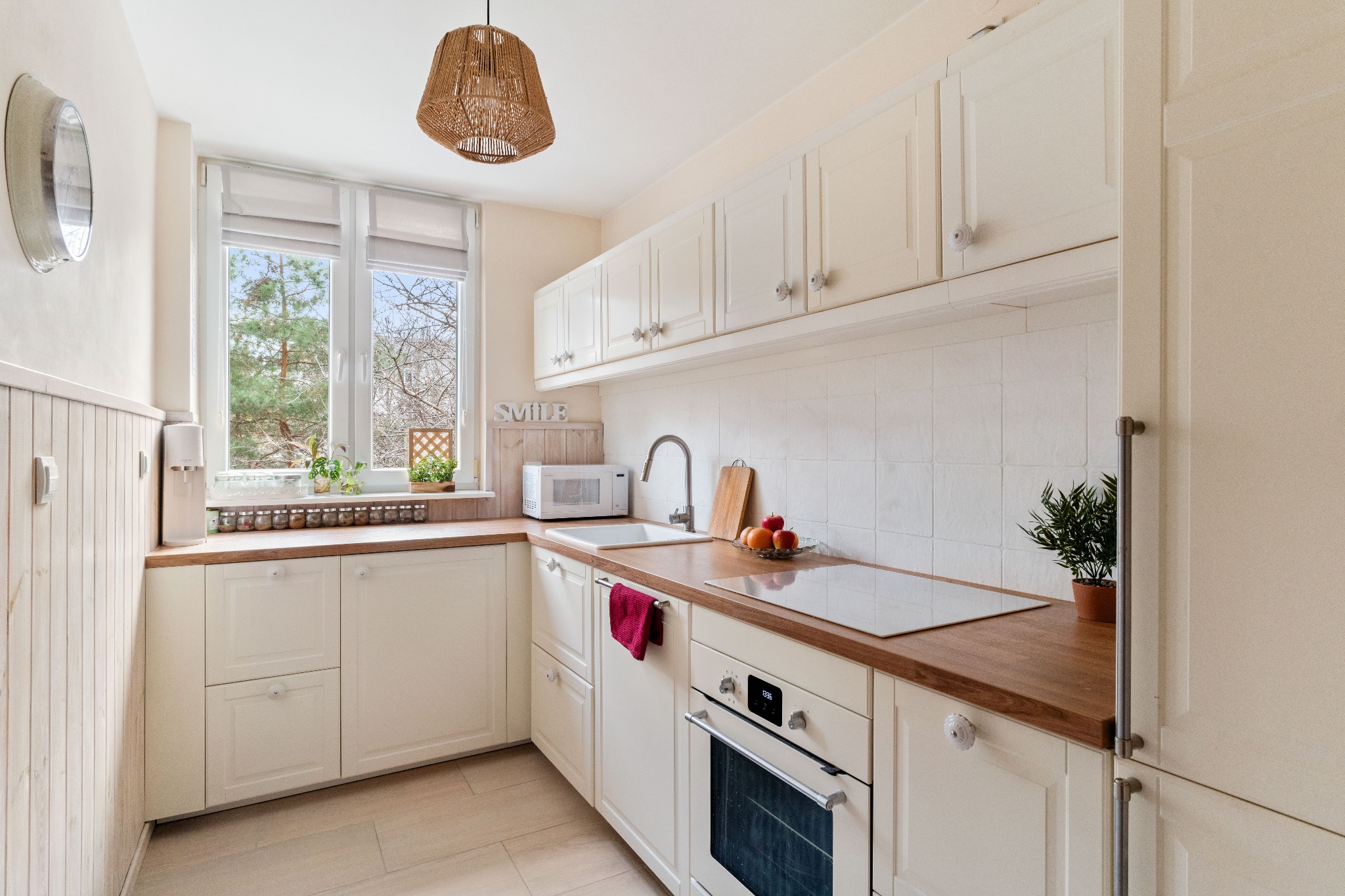 Jasna, przestronna kuchnia w stylu rustykalnym z drewnianymi blatami, białymi szafkami z ozdobnymi gałkami, widokiem na zieleń za oknem i lampą z naturalnego włókna.