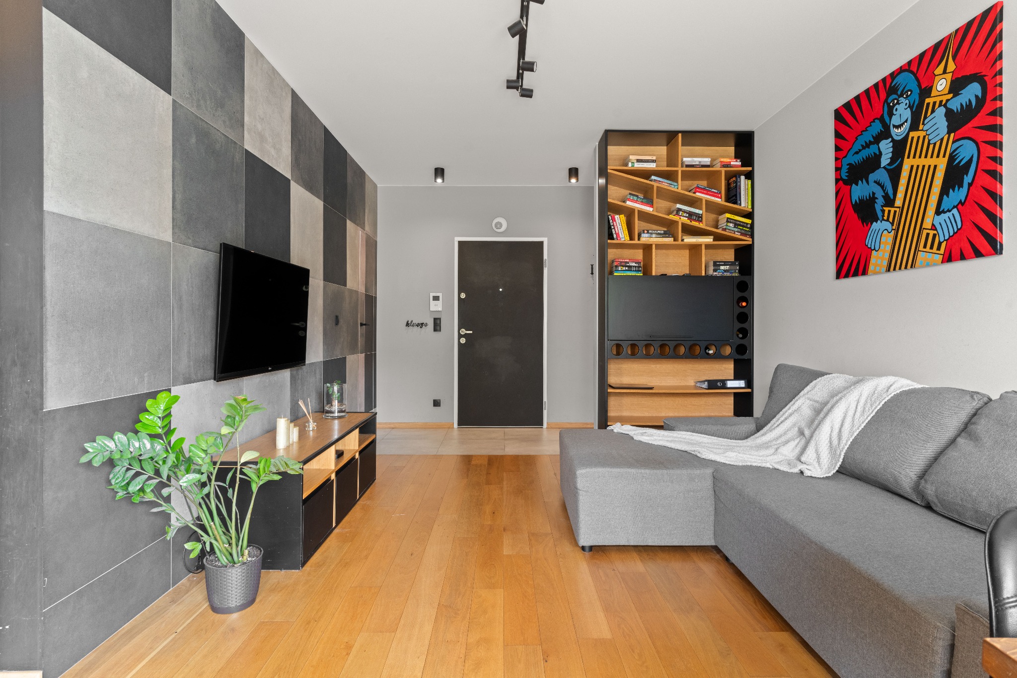 Nowoczesny salon z szarą sofą narożną, ścianą z płytek w odcieniach szarości, drewnianą podłogą i regałem z książkami, oświetlony punktowymi lampami sufitowymi i ozdobiony obrazem King Konga...