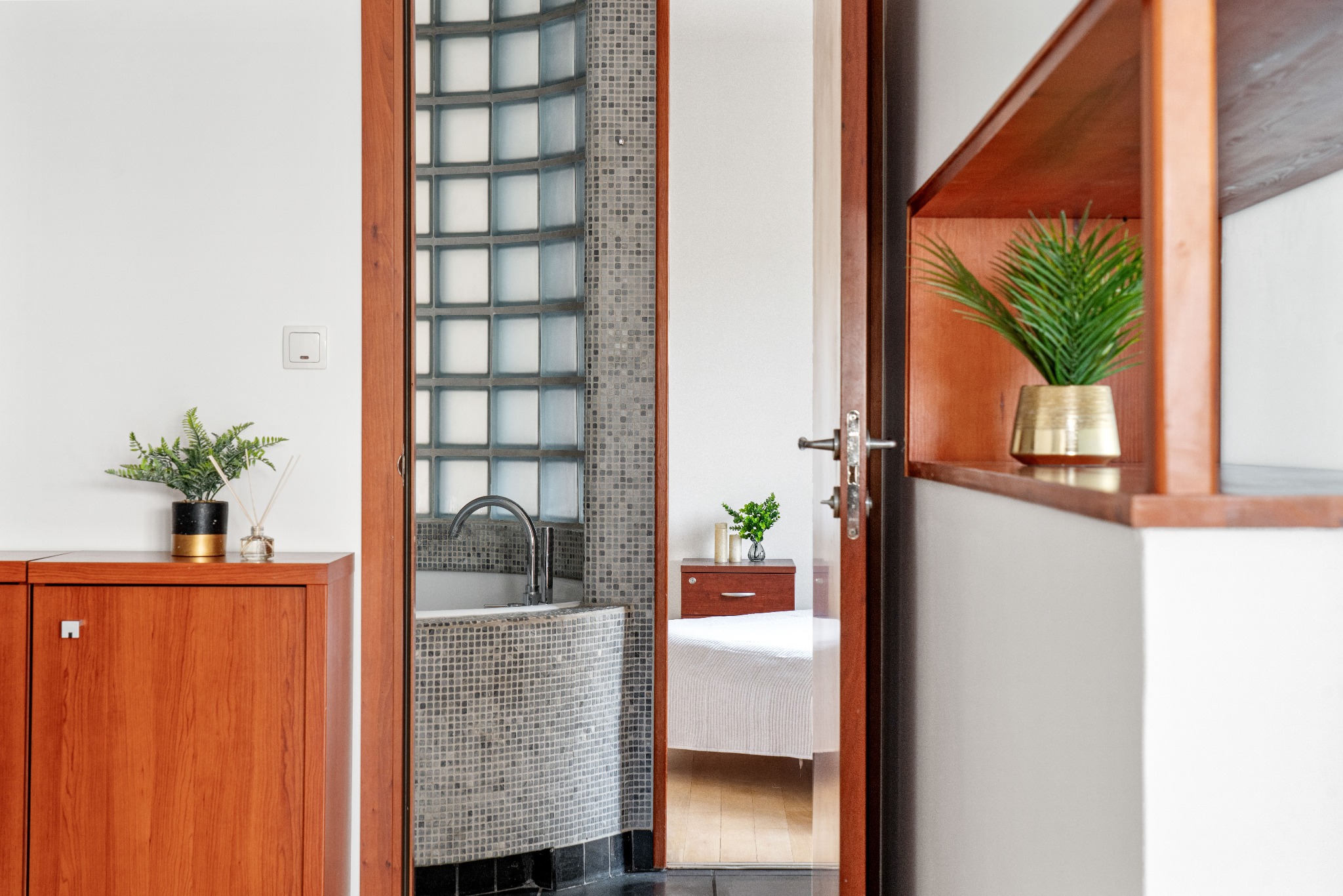 Widok na łazienkę z mozaikową wanną i szklanymi luksferami, przez otwarte drzwi widać sypialnię z drewnianymi meblami i roślinami doniczkowymi.