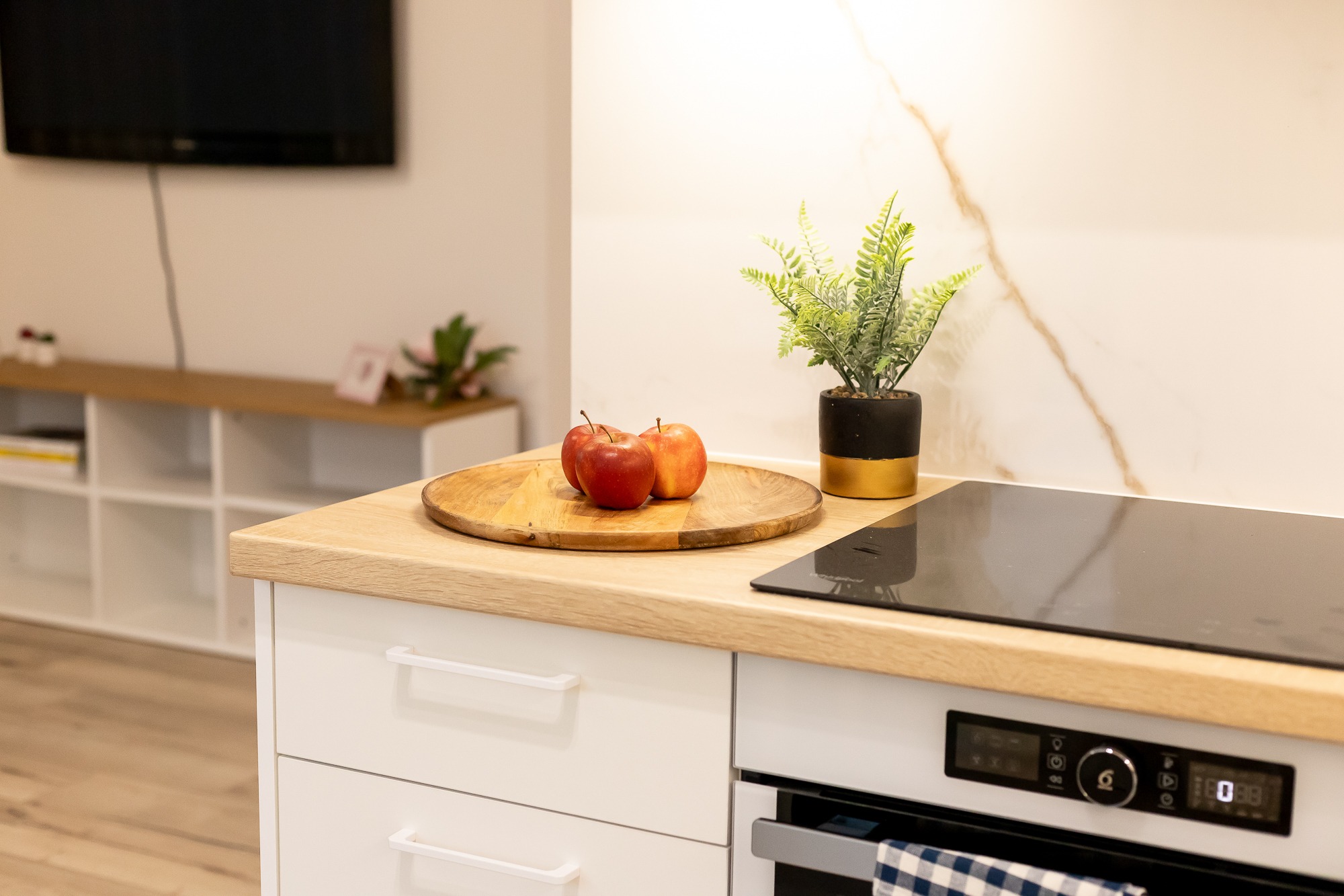 Jasna kuchnia z drewnianym blatem, na którym leżą trzy czerwone jabłka na drewnianej tacy, obok stoi roślina w czarno-złotej doniczce, w tle biała ściana z marmurowym wzorem i telewizor