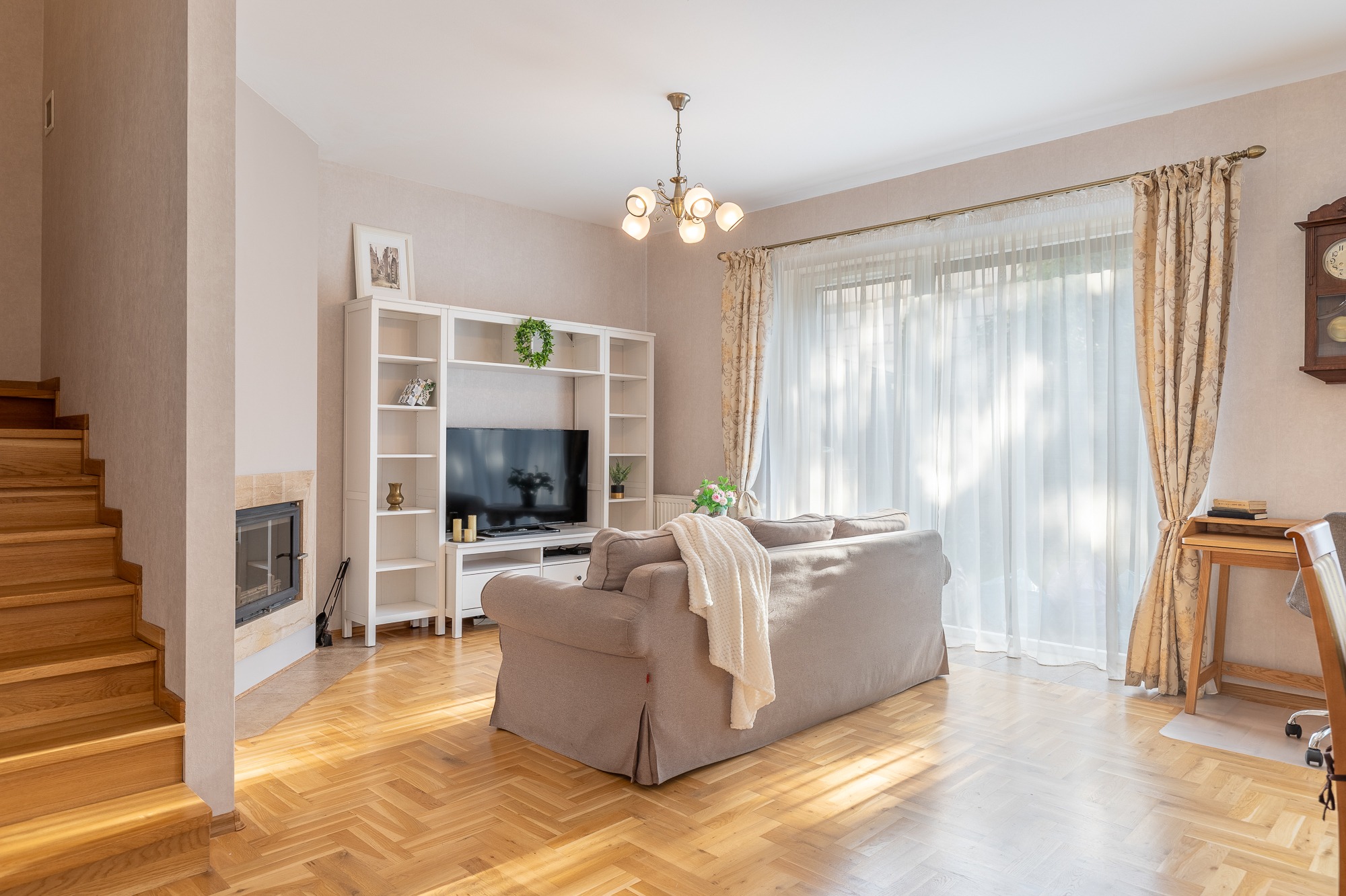 Jasne, przestronne wnętrze salonu z drewnianymi schodami, parkietem w jodełkę, beżową sofą z narzutą, białymi regałami, telewizorem, kominkiem i zasłonami w kwiatowy wzór, oświetlone światłem...