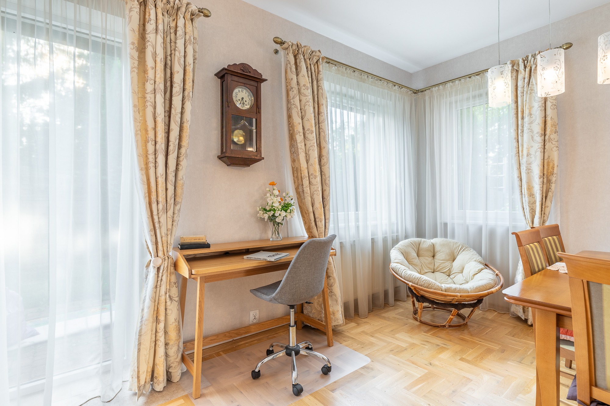 Jasne wnętrze pokoju z drewnianym biurkiem, szarym krzesłem na kółkach i okrągłym fotelem z poduszką, oświetlone naturalnym światłem wpadającym przez okna z delikatnymi, wzorzystymi zasłonami.