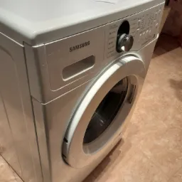 Biała pralka Samsung Ecobubble 7kg, stojąca na brązowych płytkach, z widocznym panelem sterowania i dokumentem z długopisem na górze.