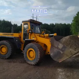 Żółta ładowarka Hanomag 55D z pełną łyżką żwiru na placu budowy, drzewa w tle.