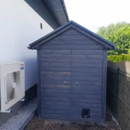 Mała, ciemnoszara drewniana budka narzędziowa z dwuspadowym dachem, stojąca obok białej ściany budynku z zamontowanym agregatem klimatyzacji i ogrodzenia z siatki.