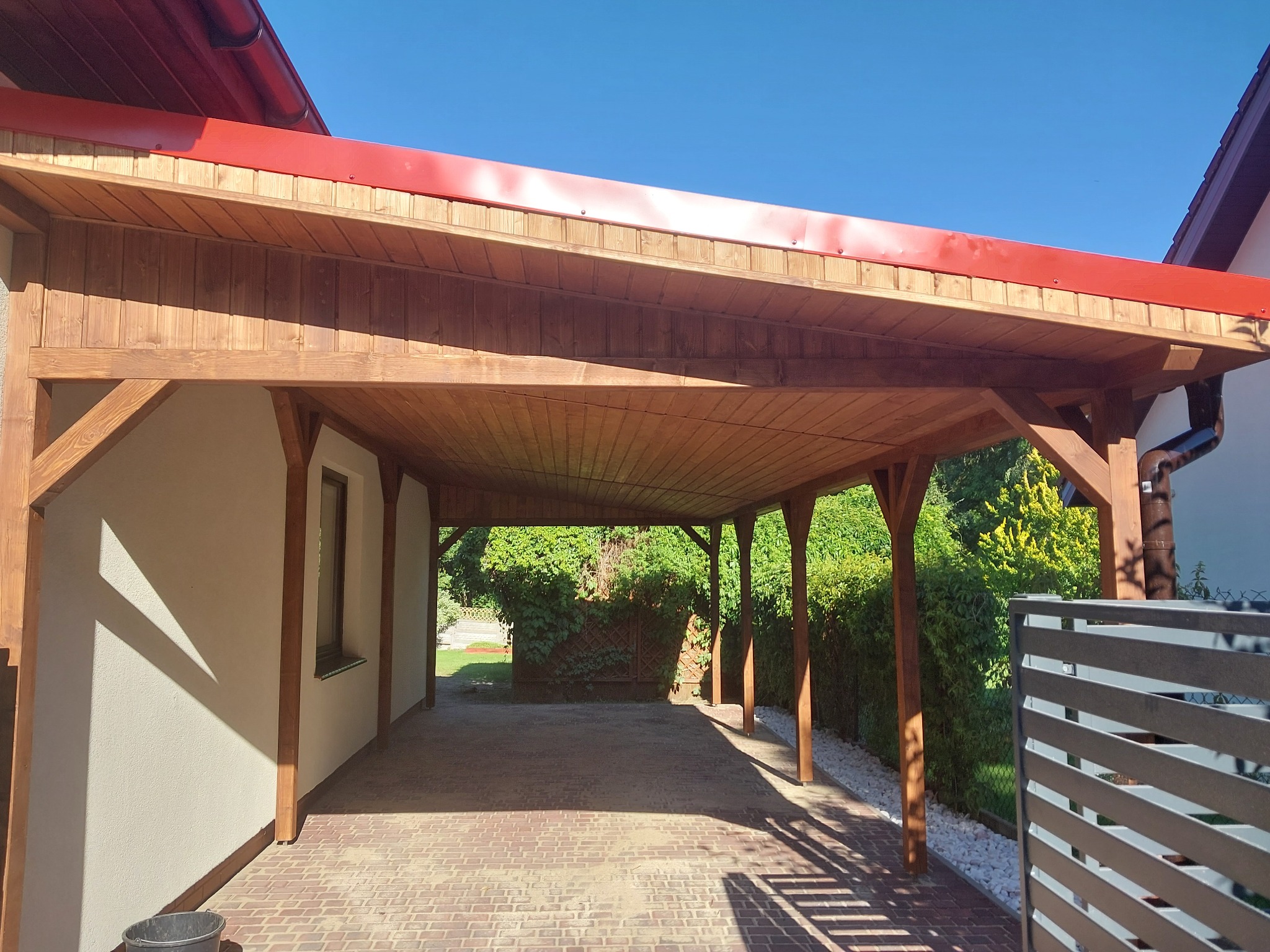 Drewniana konstrukcja z czerwonym dachem, prawdopodobnie zadaszenie tarasu lub wiata garażowa, z widoczną podłogą z kostki brukowej i fragmentem metalowego ogrodzenia po prawej stronie.