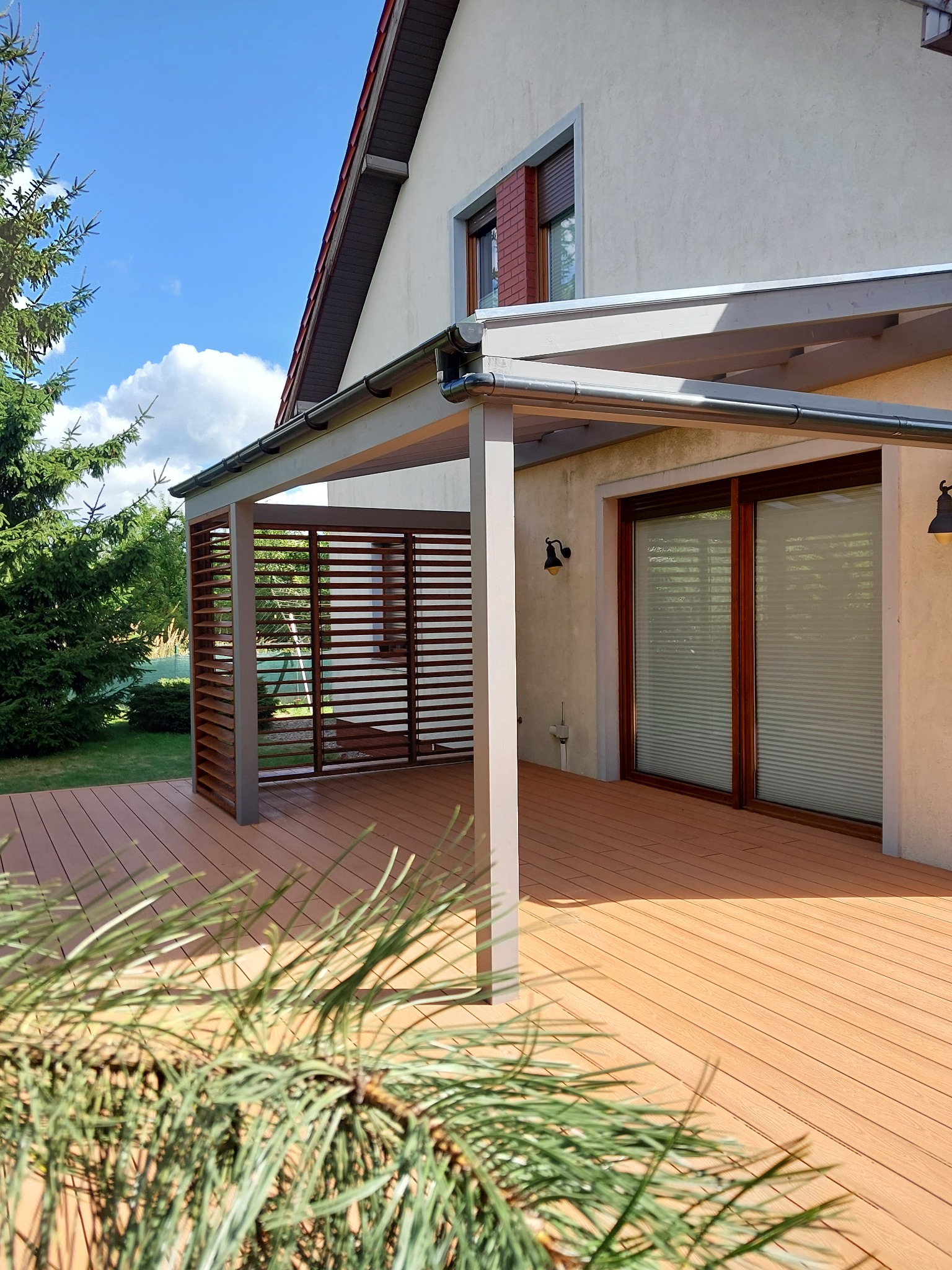 Jasny taras z kompozytu z szarą pergolą i drewnianymi lamelami, przylegający do domu z oknami z brązowymi ramami. W tle zieleń drzew i błękitne niebo.