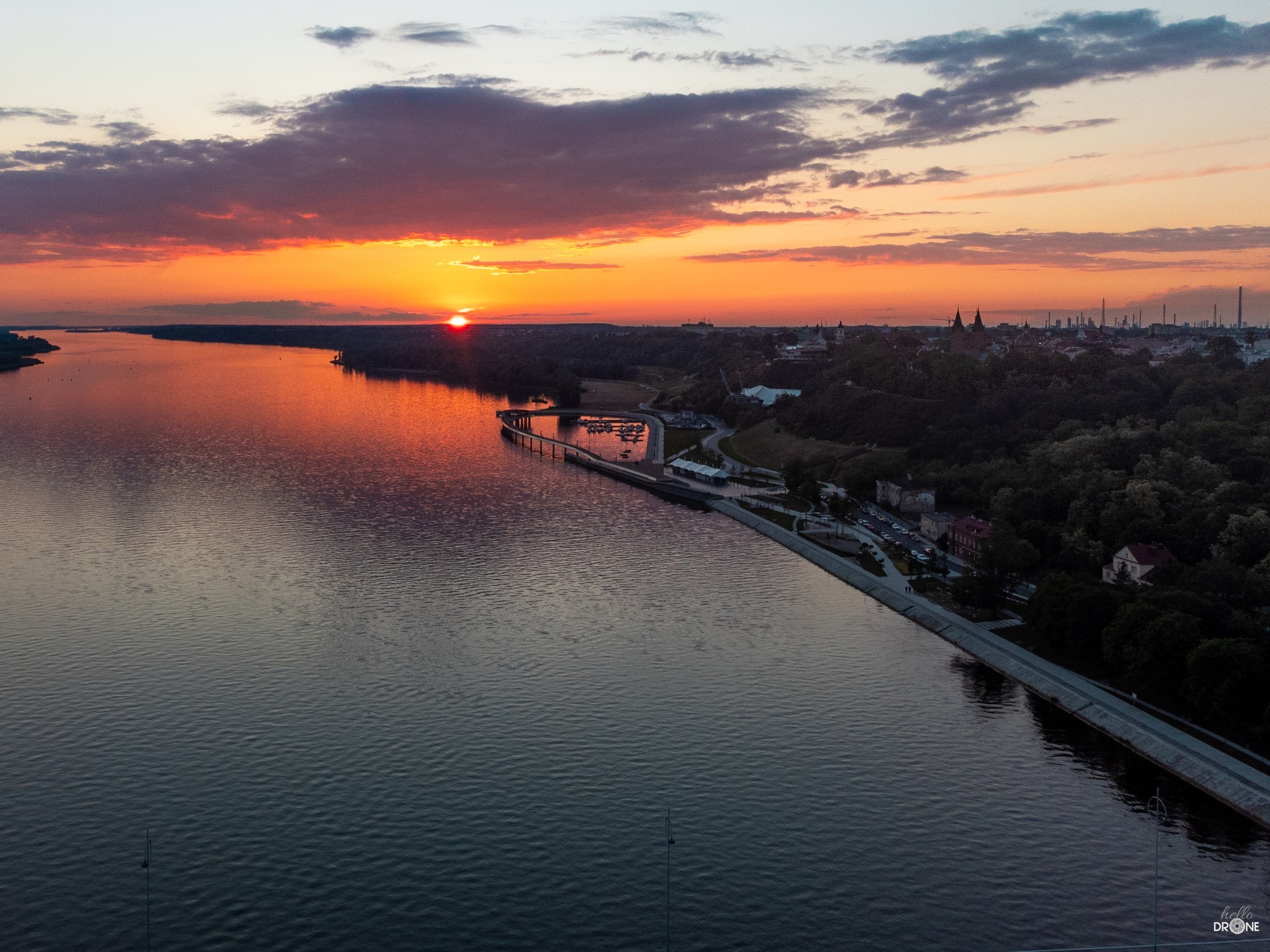 Panoramiczny widok z drona na rzekę w blasku zachodzącego słońca, z nabrzeżem, mariną i zabudowaniami miejskimi w tle, pod purpurowymi chmurami.