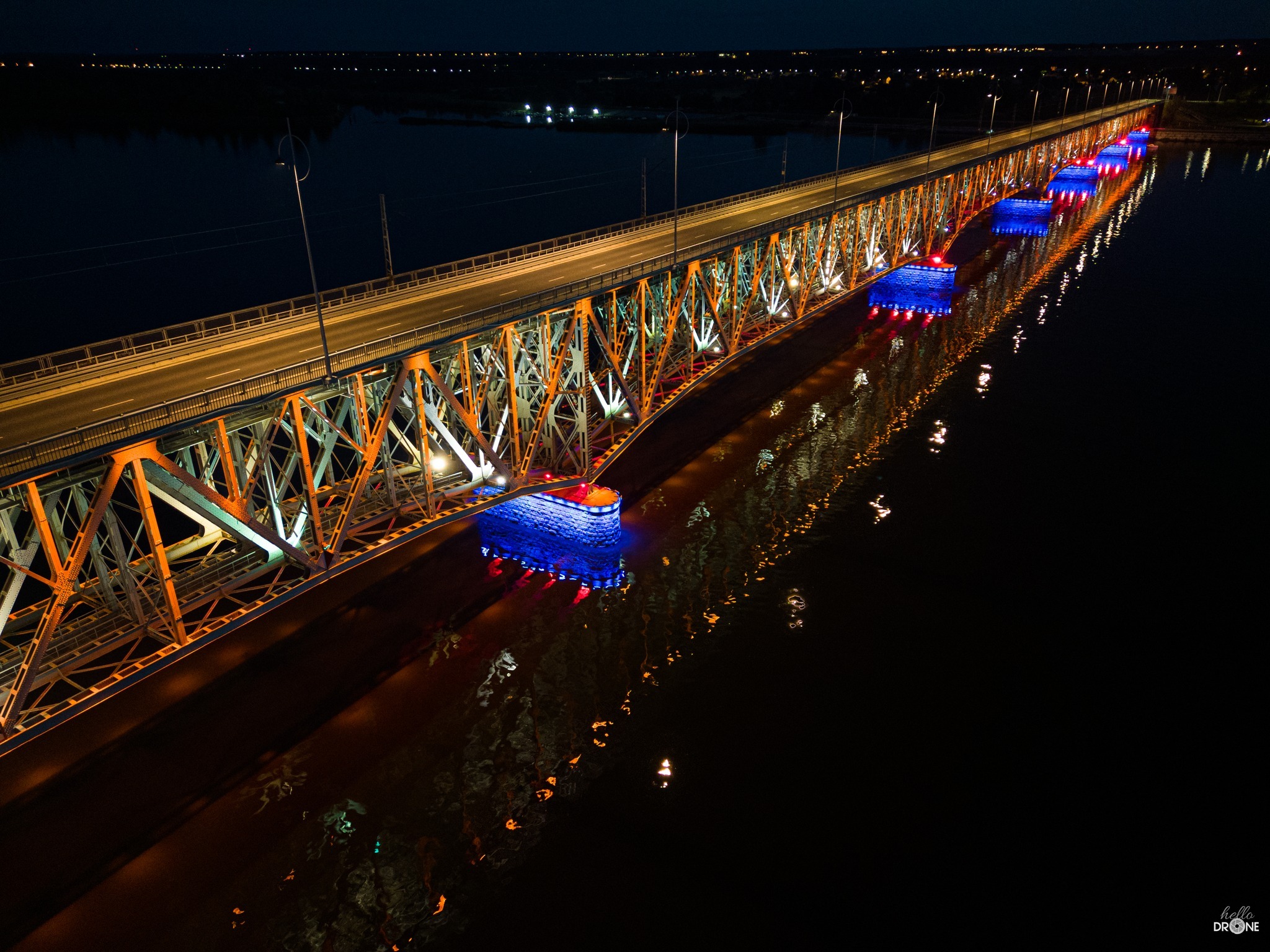 Oświetlony most stalowy nocą, odbijający się w ciemnej wodzie, widok z lotu ptaka z logo Hello Drone w rogu.