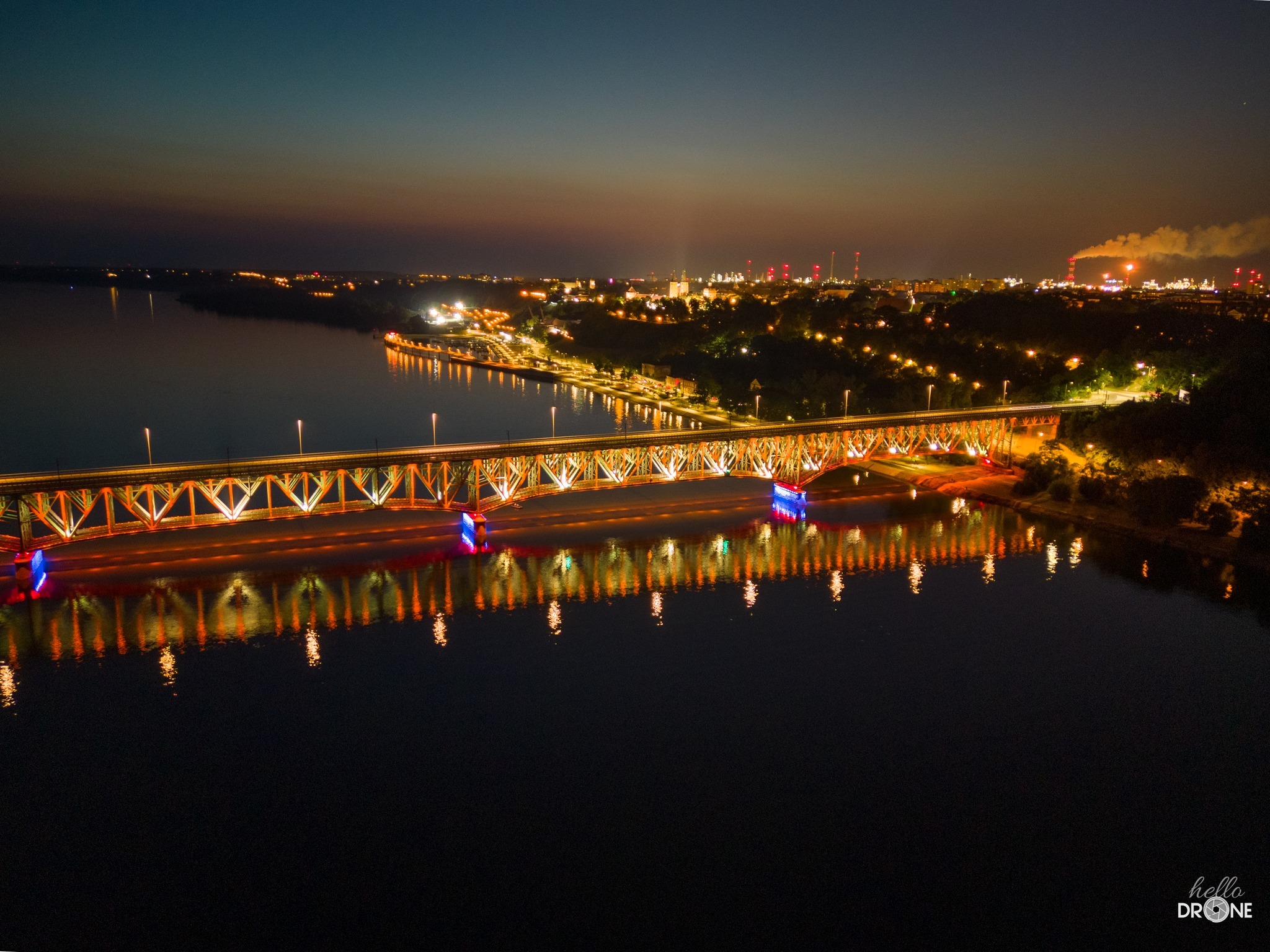 Nocny widok z drona na oświetlony most kolejowy nad rzeką, z odbiciami świateł w wodzie i panoramą miasta w tle.