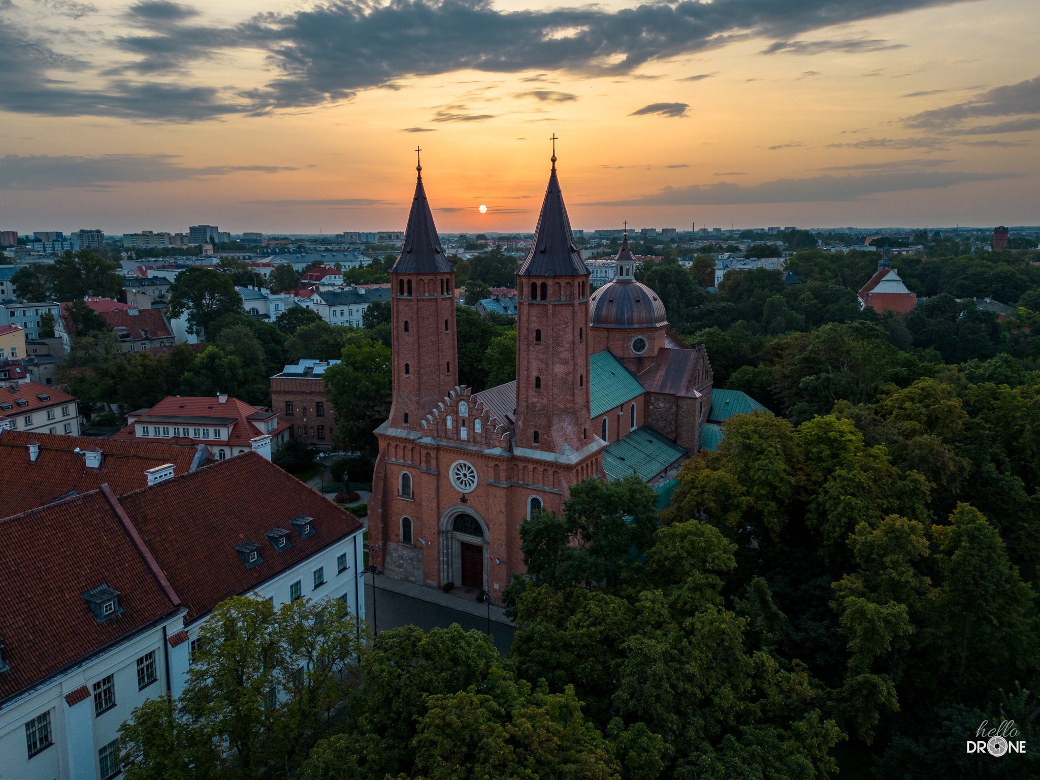 Panorama Gniezna o zachodzie słońca, widok z drona na Katedrę Gnieźnieńską z dwoma wieżami i kopułą, otoczoną zielenią drzew i zabudową miejską.