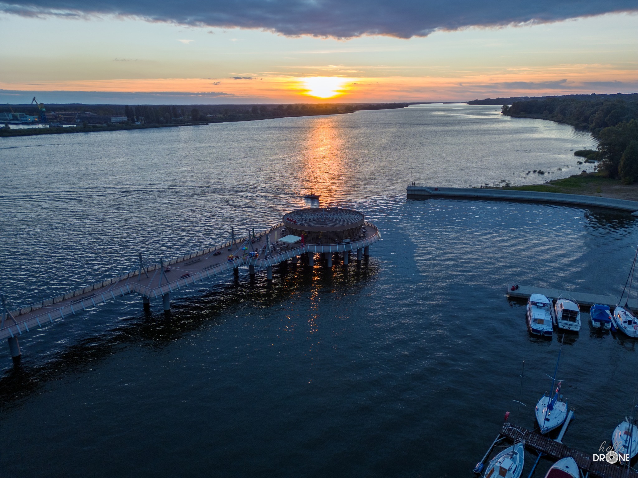Widok z drona na molo z okrągłą platformą widokową, pomost prowadzący do lądu, przystań z łodziami i zachodzące słońce nad rzeką.