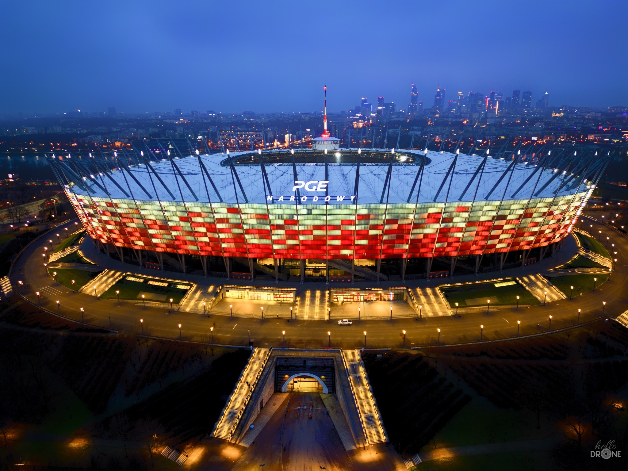 PGE Narodowy w Warszawie nocą, widok z lotu ptaka na oświetlony stadion w barwach biało-czerwonych, w tle panorama miasta z wieżowcami.