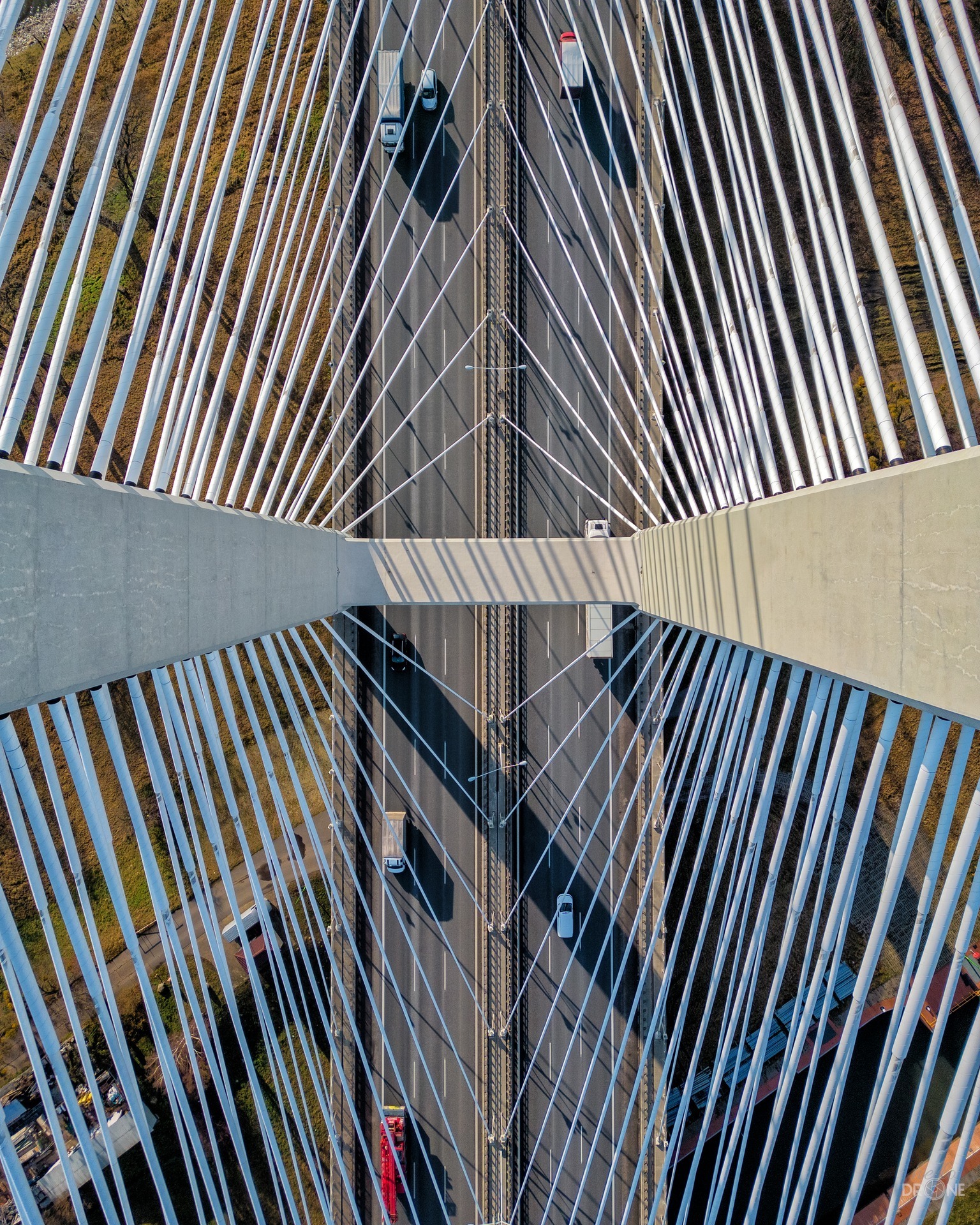 Widok z góry na most wantowy z perspektywy drona, ukazujący symetryczny układ lin i jezdnię z poruszającymi się pojazdami, otoczony zielenią.